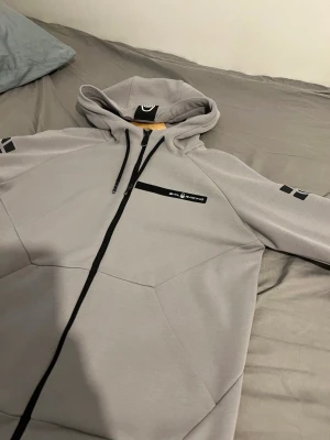 Ljusgrå zip hoodie SML Racing - Snygg ljusgrå hoodie med dragkedja från sail Racing. Hoodien har svart bröstficka med logga, svarta dragsnören i huvan och coola svarta tryck på ärmarna. Perfekt för dig som gillar streetwear och sportig stil.