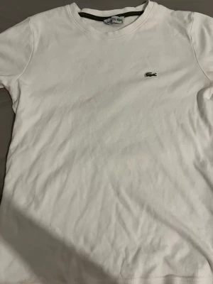  Lacoste - Klassisk vit t-shirt från Lacoste med den ikoniska krokodilloggan broderad på bröstet. T-shirten har rund halsringning och korta ärmar. Tillverkad i mjuk bomull för en skön känsla och enkel stil. Liten i storleken 