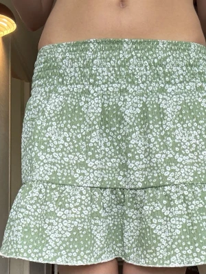 Grön blommig volangskjol - Söt kort kjol i ljusgrönt med vitt blommönster och volangkant. Kjolen har smock i midjan för en bekväm passform och är gjord i ett lätt, luftigt material som passar perfekt till varma dagar. Fin att ha som linne också💕