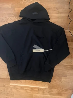 Svart hoodie Essentials Fear of God - Snygg svart hoodie från Essentials Fear of God med stor huva och känguruficka. Logga på bröstet och stor text på ryggen. Tillverkad i mjukt material som känns skönt mot huden. Perfekt för en avslappnad och trendig stil.