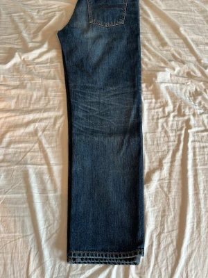 Rad Rufus jeans från nudie - Snygga mörkblå jeans från Nudie i modellen   Rad Rufus. De har en rak passform och klassisk femficksdesign. Jeansen har subtila slitningar och kontrastsömmar som ger en tidlös look. Färgen heter ”blue twisted tracks” och storleken är 30/30. Jeansen är tillverkade i denim med en autentisk känsla och passar perfekt till en avslappnad stil.
