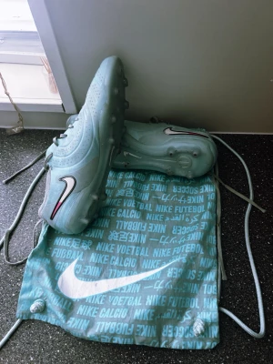 Ljusblå Nike fotbollsskor - Säljer ett par ljusblå Nike fotbollsskor med klassisk svart och vit Swoosh-logga och röda detaljer. Skorna har snörning och dobbar för bra grepp på planen. Medföljer en matchande Nike-påse med tryck. Perfekta för dig som vill sticka ut på fotbollsplanen.