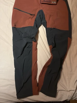 Tvåfärgade friluftsbyxor Haglöfs, Rugged Flex Pants - Säljer ett par snygga tvåfärgade friluftsbyxor från Haglöfs i dammodell, storlek 38. Byxorna är i rostbrunt/vinröda (svårt att få exakt färg på foto) och svart, har dragkedjeficka framtill och förstärkta partier på knän och bak. Resår i midjan och kardborreknäppning runt ankel. Perfekta för uteliv och äventyr.