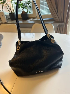 Svart handväska från Michael Kors - Stilren svart handväska från Michael Kors i mjukt, strukturerat läder. Väskan har två långa axelband, guldfärgade metalldetaljer och flera fack inuti, inklusive ett med dragkedja. Perfekt för dig som vill ha både stil och funktion.