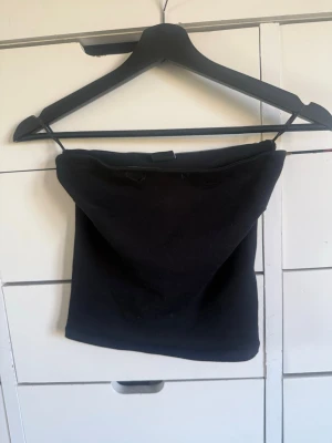Svart tubtopp i bomull - En enkel svart tubtopp i mjukt bomullsmaterial. Toppen är axelbandslös och har en klassisk bandeau-form som sitter tight runt överkroppen. Perfekt att styla med jeans eller kjol för en clean look.