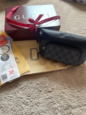Gucci axelväska i grå monogram - Snygg axelväska från Gucci i grått med klassiskt GG-monogram. Väskan har en kompakt form, svart dragkedja och en praktisk ytterficka framtill. Tillbehör som dustbag, box och certifikat medföljer. Perfekt för dig som vill ha en stilren och lyxig accessoar.