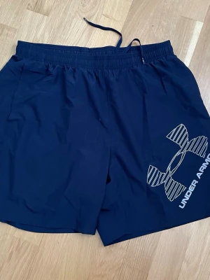 Marinblå träningsshorts Under Armour - Snygga marinblå träningsshorts från Under Armour med stort logotryck på ena benet. Shortsen har elastisk midja med snörning och är tillverkade i ett lätt och snabbtorkande material, perfekta för gymmet eller löprundan.