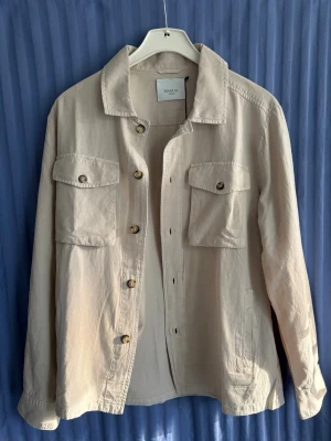 Beige overshirt från Marzia. Helt ny - Stilren beige overshirt från Marzia med klassisk krage och två stora bröstfickor med knappar. Overshirten har långa ärmar och knäppning framtill. Perfekt att slänga över en t-shirt för en avslappnad look.