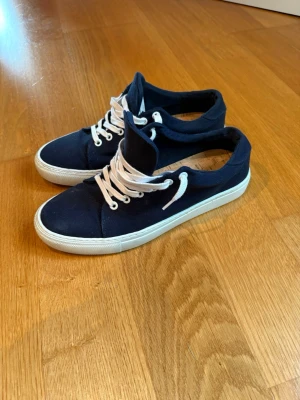 Mörkblå canvas sneakers med vita snören - Snygga mörkblå sneakers i canvas med vita snören och vit sula. Klassisk låg modell som passar till det mesta och ger en clean look. Perfekta för dig som gillar enkel och stilren design.