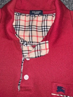Röd kortärmad topp från Burberry - Snygg röd topp från Burberry med klassiskt rutigt mönster i kragen och på fickan. Kortärmad modell med knappslå och liten bröstficka med broderad logga. Perfekt för dig som gillar stilrena detaljer och ikonisk design.