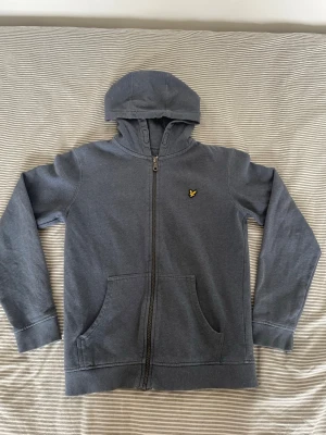 Blå zip-hoodie med logga - Snygg blå hoodie med dragkedja och huva. Har en gul broderad logga på bröstet och två stora fickor framtill. Tillverkad i mjukt sweatshirtmaterial och har ribbade muddar vid ärmar och nederkant. Perfekt för en avslappnad stil.