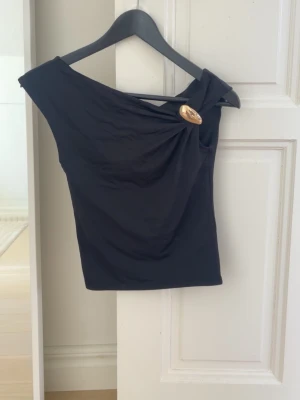Svart oneshoulder topp med detalj - Snygg svart oneshoulder topp med en  guldfärgad knappdetalj vid axeln.