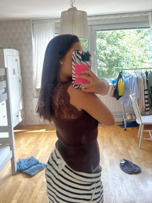 Mörkbrun ribbad spetslinnetopp - Säljer en mörkbrun ribbad linnetopp med fina spetsdetaljer upptill och vid axlarna. Toppen är ärmlös och har en kroppsnära passform som framhäver figuren. Perfekt för dig som gillar en stilren men ändå söt look.