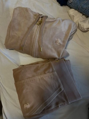 Beige velour set från Juicy Couture - Säljer en beige velour hoodie från Juicy Couture med guldig dragkedja, huva och guldfärgade detaljer. Hoodien har broderad logga på bröstet och är supermjuk och skön. Perfekt för en avslappnad och trendig look. Koftanr i Stl Xs och byxorna i stl S. Köpt i butik på raglady, har kvittobevis. Har endast handtvättas, och använd 3 gånger.