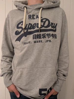Grå Superdry hoodie med tryck - Snygg grå hoodie från Superdry med stort mörkblått tryck på bröstet och japanska tecken. Hoodien har huva med vita dragsnören och en stor magficka. Tillverkad i mjukt sweatshirtmaterial, perfekt för chill dagar.