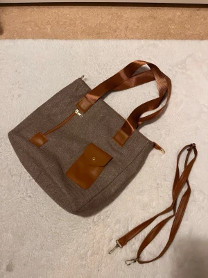 Brun axelväska med detaljer - Snygg brun axelväska i tyg med bruna detaljer i konstläder. Väskan har både korta och avtagbar lång axelrem, en framficka med knapp och en snedställd dragkedja. Perfekt för dig som vill ha en stilren och praktisk väska med plats för det viktigaste.