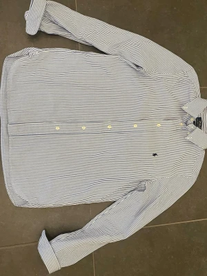 Randig skjorta från Polo Ralph Lauren - Klassisk skjorta från Polo Ralph Lauren med blå och vita ränder. Skjortan har lång ärm, knappar framtill och den ikoniska broderade loggan på bröstet. Perfekt till jeans eller chinos för en clean och stilren look.                              slim fit S i storlek.       För liten för mig men passar bra från 165-170cm 