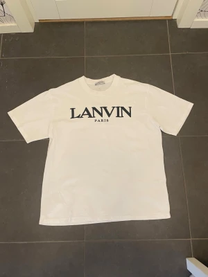 Vit t-shirt  - Säljer en clean vit t-shirt. Med en stor svart logga på bröstet. Klassisk rund halsringning och korta ärmar. Perfekt basic-plagg med exklusiv känsla och skön passform i mjuk bomull. 