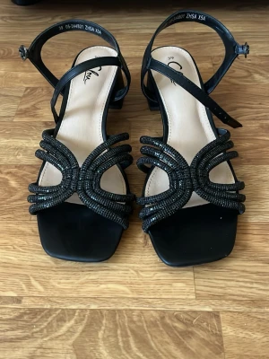 Svarta sandaler med blockklack - Stilrena svarta sandaler med fyrkantig tå och låg blockklack. Remmarna framtill är dekorerade med svarta pärlor i ett unikt mönster och har justerbar ankelrem med spänne. Perfekta för dig som vill ha en trendig och bekväm look till sommaren.