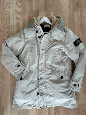 Beige vinterparkas från Stone Island - Snygg beige vinterparkas från Stone Island med klassisk patch på ärmen. Jackan har stora fickor framtill, knappar och dragkedja samt en mysig krage med detaljer och foder i teddy. Perfekt för kyliga dagar. Modellen är rak och jackan är väldigt sparsamt använd pga fel storlek. Nypris cirka 14-15 000 SEK. 