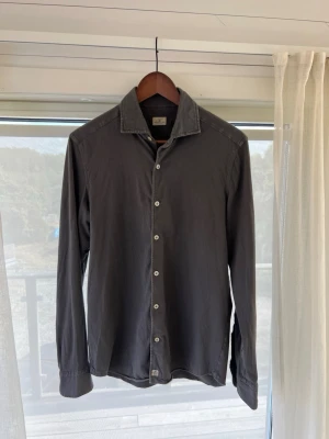 Sonrisa - Säljer en stilren mörkgrå skjorta från Sonrisa med klassisk krage och vita knappar. Skjortan har lång ärm och är tillverkad i mjuk bomull, perfekt för en clean och avslappnad look.