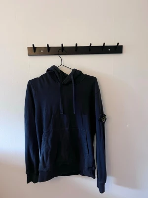 Svart hoodie från Stone Island - Snygg hoodie från Stone Island med klassisk patch på vänster ärm. Hoodien har huva och en stor magficka framtill. Tillverkad i mjukt bomullsmaterial och har ribbade muddar vid ärmslut och nederkant. Perfekt för en avslappnad streetwear-look.