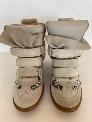 Beige höga sneakers med kardborre - Unika beige sneakers med högt skaft och tre breda kardborreband. Skorna har detaljer i mocka och tyg med svartvitt mönster på sidorna och framtill. Sulan är i gummi och designen är chunky och trendig, perfekt för dig som vill sticka ut.
