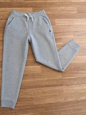 Ralph Lauren byxor - Säljer ett par klassiska mjukisbyxor från Polo Ralph Lauren i ljusgrå färg med broderad logga. Byxorna har en bekväm passform med resår och dragsko i midjan samt muddar vid anklarna – perfekta för både hemmabruk och vardag. • Märke: Polo Ralph Lauren • Färg: Grå • Storlek: L (14-16) • Skick: Mycket fint skick, sparsamt använda😁 • Detaljer: Dragsko i midjan, sidofickor och broderad logga!