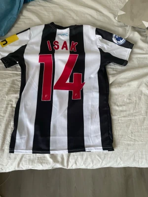Newcastle United matchtröja Isak 14 - Säljer en svartvit randig Newcastle United matchtröja med Isak och nummer 14 på ryggen. Tröjan har klubbmärke på bröstet, sponsortryck i blått och detaljer i gult på ärmen. Tillverkad i lätt och ventilerande material, perfekt för fotboll eller träning.