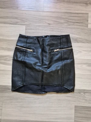 Svart kort kjol i skinnimitation - Snygg svart kort kjol i skinnimitation med två dekorativa dragkedjor framtill. Kjolen har en tight passform och en lätt glansig yta som ger en cool look. Perfekt att matcha med en t-shirt eller stickad tröja.