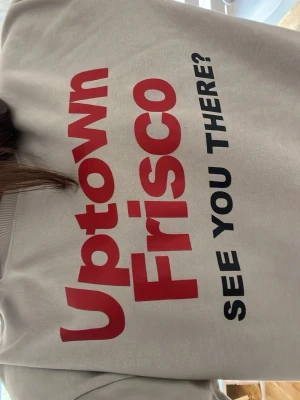 Grå hoodie med Uptown Frisco-tryck - Säljer en grå hoodie med stort rött och svart tryck på framsidan där det står 'Uptown Frisco SEE YOU THERE?'. Hoodien har en avslappnad passform och ribbade muddar vid ärmar och nederkant. Perfekt för en chill och trendig look.