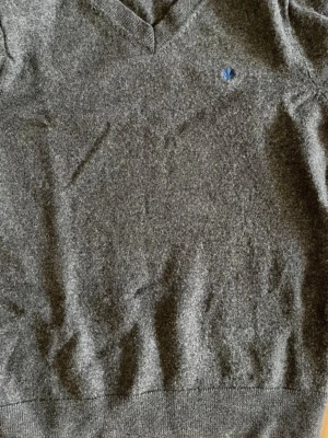 Grå V-ringad tröja från Morris - Mjuk och stilren grå V-ringad tröja från Morris i Merino ull. Tröjan har en liten blå broderad detalj på bröstet och ribbade muddar vid hals, ärmslut och nederkant. Perfekt för lager på lager och enkel att matcha med olika outfits.