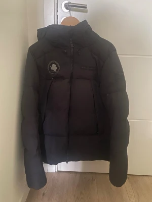 Sail racing vinterjacka - Säljer en svart pufferjacka med huva och dragkedja framtill. Jackan har två bröstpatchar och en patch på ärmen för en cool look. Fylld med vaddering för extra värme och har en klassisk pufferdesign med breda paneler. Jackan är i gott skick!