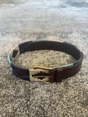 Brunt och blått mönstrat bälte - Snyggt bälte i brunt skinn med detaljer i blått, beige och mörkblått. Bältet har ett unikt geometriskt mönster och ett klassiskt metallspänne. Perfekt för att lyfta din outfit med lite extra färg och stil.