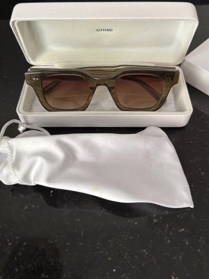 Olivgröna solglasögon från CHIMI - Snygga solglasögon från CHIMI i olivgrönt transparent acetat med fyrkantig form och bruna tonade glas. Kommer med vit hård förvaringsbox och mjukt fodral. Perfekta för dig som vill ha en stilren och modern look.