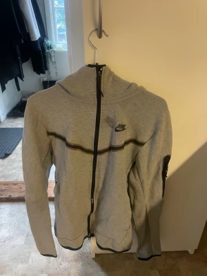 Grå zip hoodie från Nike - Snygg grå hoodie från Nike med hel dragkedja framtill och svart Nike-logga på bröstet. Hoodien har en huva, svarta detaljer längs dragkedjan och ärmslut samt en ficka på ena ärmen. Perfekt för en sportig och avslappnad stil.