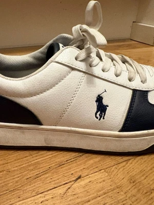 Vita och marinblå Polo Ralph Lauren sneakers - Snygga sneakers i vitt och marinblått från Polo Ralph Lauren med klassisk logga på sidan. Skorna har vita snören, låg profil och ovandel i läderimitation. Perfekta för en clean och stilren look.