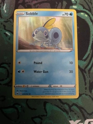 Pokémonkort Sobble - Säljer ett Pokémonkort med Sobble, en vatten-Pokémon från Sword & Shield-serien. Kortet har blå och gul ram, med Sobble illustrerad i mitten. Kortet har attackerna Pound och Water Gun samt HP 70. Perfekt för samlare eller Pokémonfans.