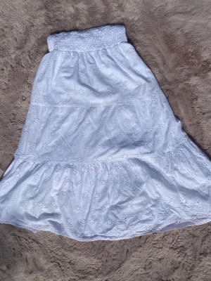 Vit långkjol med broderade detaljer - Söt vit långkjol i bomull med broderade blommor och bred, smockad midja. Kjolen har volang nedtill och ett luftigt, somrigt uttryck. Perfekt för dig som gillar romantisk och avslappnad stil.