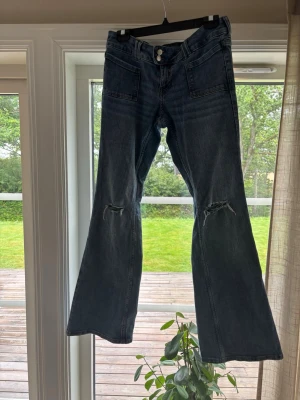 Mörkblå bootcut jeans med slitningar - Säljer ett par mörkblå bootcut jeans med slitningar över knäna och stora framfickor. Jeansen har utsvängda ben och klassiska bakfickor. Materialet är denim i bomull och de har en cool vintage vibe.