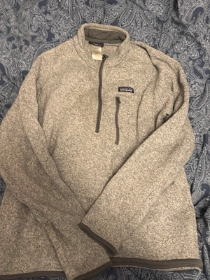 Grå fleece-tröja från Patagonia - Mysig gråmelerad fleece-tröja från Patagonia med halv dragkedja och bröstficka med dragkedja. Tröjan har hög krage och detaljer i mörkgrått vid muddar och nederkant. Perfekt för lager-på-lager och outdoor.