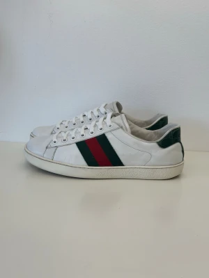 Vits Gucci ace sneakers - Snygga vita sneakers i läder med klassiska gröna och röda sidoremsor samt gröna detaljer vid hälen. Skorna har vit sula och vita skosnören. Perfekta för dig som vill ha en stilren och trendig look.