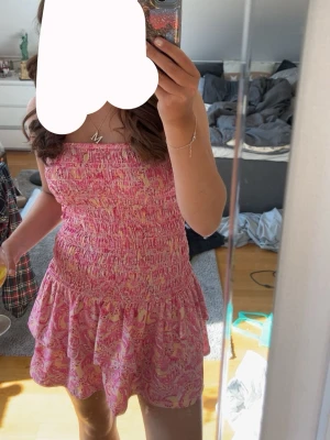 Rosa kortklänning med volang - Supersöt rosa kortklänning med smockad överdel och volang i nederkant. Klänningen har ett blommigt mönster i rosa och ljusa toner samt är ärmlös med rak urringning. Perfekt för varma dagar och ger en fräsch sommarkänsla. Använd cirka 3-4 gånger 