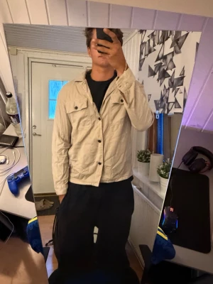 Beige overshirt med fickor - Säljer en beige overshirt med två bröstfickor och knappar framtill. Overshirten har klassisk krage och långa ärmar med knapp vid ärmslut. Materialet ser ut att vara bomull eller bomullsblandning, perfekt för lager på lager och en avslappnad stil.