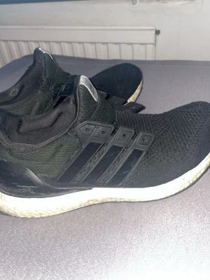 Svarta Adidas Ultraboost sneakers - Svarta Adidas Ultraboost sneakers med stickad ovandel och vita Boost-sulor. Skorna har de klassiska tre svarta ränderna på sidan och en flexibel, låg siluett. Perfekta för dig som gillar sportig och stilren design.