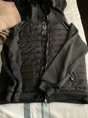 Svart hoodie med quiltad front - Säljer en svart hoodie med quiltad frontpanel och dragkedja. Tröjan har huva, långa ärmar och fickor med dragkedja. Perfekt för dig som gillar en sportig och stilren look.