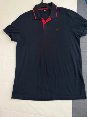 Marinblå pikétröja från BOSS - Snygg marinblå pikétröja från BOSS med röda detaljer på krage och knappslå. Klassisk passform och korta ärmar, perfekt till både jeans och shorts. BOSS-logga broderad på bröstet för en clean look.