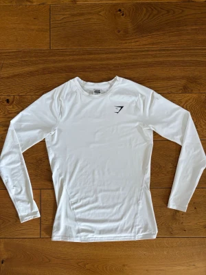 Vit långärmad träningströja Gymshark - Säljer en vit compression shirt från Gymshark. Tröjan är tight fit med rund hals och har en diskret svart Gymshark-logga på bröstet. Passar perfekt för gymmet.