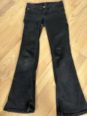 Svarta bootcut jeans med rosa detalj - Snygga svarta bootcut jeans med rosa patch på bakfickan. Jeansen har klassisk femficksdesign, knappgylf och utsvängda ben. Materialet ser ut att vara denim med lite stretch för skön passform. Perfekta för en trendig och avslappnad look. Väldigt säker på att det är stl är 27 midjan och 30 i benen med det står ingenstans!