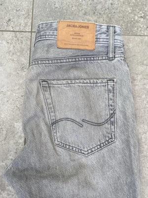 Grå jeans från Jack & Jones - Snygga grå jeans från Jack & Jones med klassisk femficksdesign och raka ben. Jeansen har en ljus tvättad look och är tillverkade i denim med tydliga sömmar och läderpatch bak i midjan. Perfekta för en avslappnad och trendig stil.